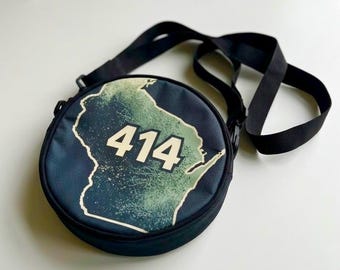 May include: A round black crossbody bag featuring a green and white graphic of the state of Wisconsin with the number 414 printed in large white font across the center. The bag includes an adjustable black strap attached to the sides.