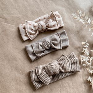 May include: Three knotted headbands in neutral colors. One headband is a light brown with a waffle texture, one is a light gray, and one is a light beige with a small floral pattern.