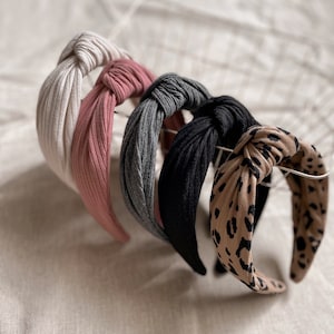 May include: Four knotted headbands in different colors and patterns. The headbands are made of a soft, stretchy fabric. One headband is white, one is pink, one is gray, and one is black with a leopard print.