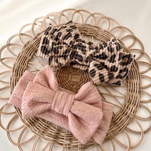 May include: Two fabric headbands with large bow details. One has a leopard print pattern in brown and black. The other is a solid pale pink. Both are displayed on a woven, circular placemat, presented in a neutral, natural colour.