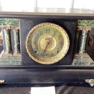 May include: Antique black mantel clock with a round, gold-toned clock face. The clock features green marble accents and decorative columns. The base has ornate feet. The clock's face shows the time.