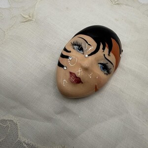 May include: A ceramic brooch shaped like a stylized face with black hair, blue eyes, and red lips. The face features black painted markings on the side and is decorated with small, clear rhinestones embedded on the forehead and cheek area.