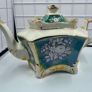 May include: A vintage porcelain teapot with a square base, featuring a white and teal body with gold trim. The teapot has a floral design and a green lid with gold accents. The handle and spout are also trimmed in gold.