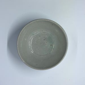 May include: A handmade, light gray ceramic bowl with a slightly textured surface. The bowl has a wide, shallow design and a small, stamped marking on the interior base. The bowl is suitable for serving food.