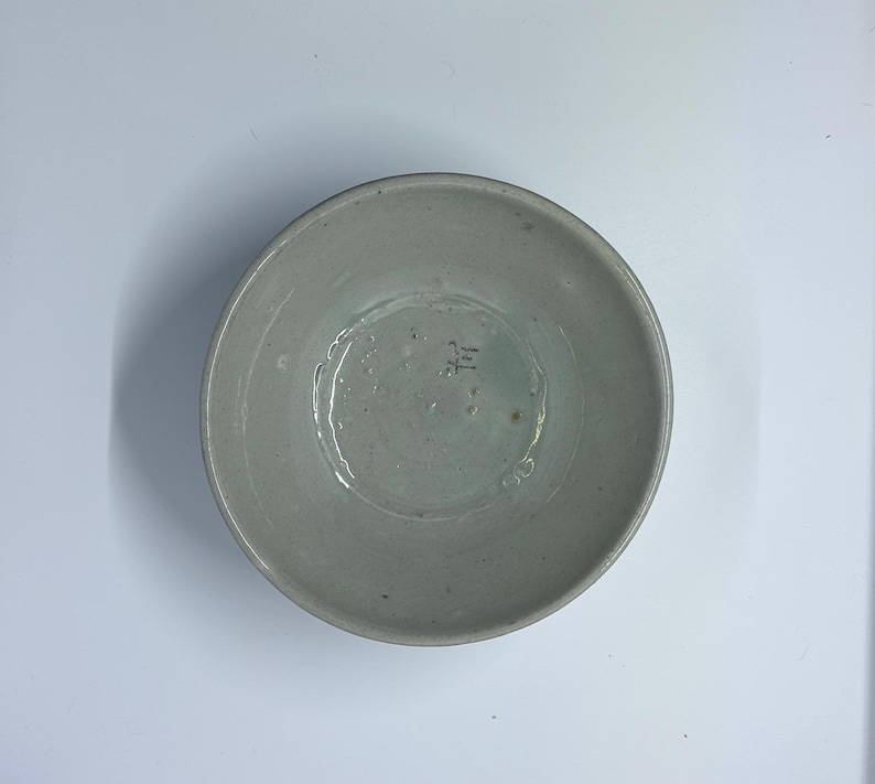 May include: A handmade, light gray ceramic bowl with a slightly textured surface. The bowl has a wide, shallow design and a small, stamped marking on the interior base. The bowl is suitable for serving food.