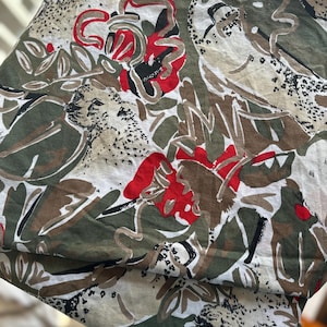 May include: A folded fabric with a botanical print featuring red, green, brown, and black floral and leaf designs. The fabric has a white background with a camouflage-like pattern.