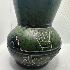 May include: Green ceramic vase with a bulbous body and a flared top. The vase features etched designs in white and black, including geometric and stylized motifs. The surface has a speckled texture, adding visual interest to the handcrafted pottery.