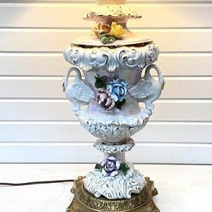 May include: A vintage ceramic table lamp with an ornate design. The lamp features a white and pink urn-shaped base decorated with swan figures, floral accents, and a gold-toned metal base. The lamp has a decorative top with a floral design.