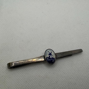 Pode incluir: Uma presilha de gravata prateada com um design oval em esmalte azul e branco. O esmalte apresenta uma cena de moinho de vento. A presilha é retangular e tem um fecho na parte de trás.
