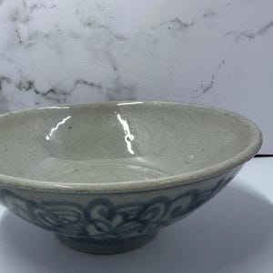 May include: A light gray ceramic bowl with a wide, shallow design. The bowl features a decorative blue floral pattern around the base. The interior is smooth and reflective, suggesting a glazed finish. This bowl is suitable for serving food.