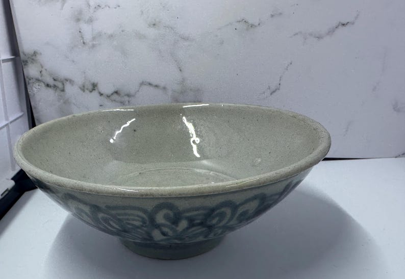 May include: A light gray ceramic bowl with a wide, shallow design. The bowl features a decorative blue floral pattern around the base. The interior is smooth and reflective, suggesting a glazed finish. This bowl is suitable for serving food.