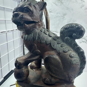 May include: A bronze-colored lamp base featuring a detailed sculpture of a mythical guardian lion. The lion is seated, holding a ball, with intricate fur and mane details. The lamp has a brown metal stem and a decorative top.