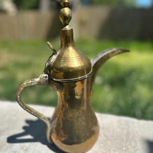 May include: A vintage brass coffee pot with a curved spout and handle. The pot has a bulbous body and a decorative finial on the lid. The brass has a warm, aged patina, suggesting its use over time. The pot is likely used for serving coffee or tea.