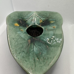 May include: A green ceramic flower frog shaped like a leaf. The frog has a central hole with a metal pin holder for arranging flowers. The leaf design has visible veins and a glossy finish with some imperfections.