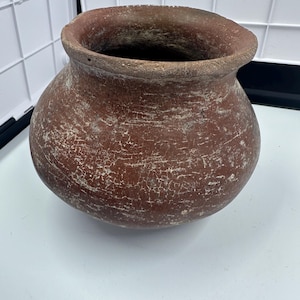 Könnte beinhalten: Ein runder, rotbrauner Steinguttopf mit einer weiten Öffnung. Der Topf hat einen bauchigen Körper und einen leicht ausgestellten Rand. Die Oberfläche ist strukturiert mit sichtbaren Rissen und einem verblassten, gealterten Aussehen. Der Topf ist wahrscheinlich ein antikes oder Vintage-Objekt.