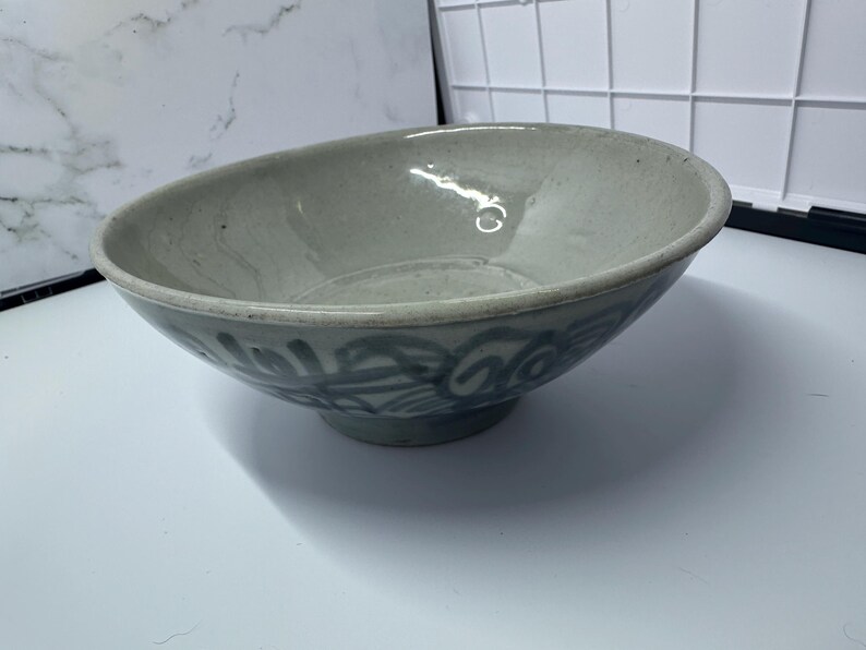 May include: A light gray ceramic bowl with a slightly angled rim. The bowl has a footed base and is decorated with a blue, swirling pattern around the exterior. The bowl is likely used for serving food.