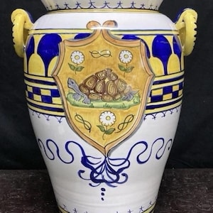 May include: A large, ornate ceramic vase with a white base, adorned with blue and yellow accents. The vase features a central shield design with a turtle illustration. Yellow handles and a blue and yellow rim complete the design.