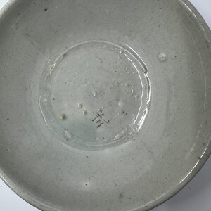 May include: A light gray ceramic bowl with a slightly textured surface. The bowl has a wide, shallow design with a rounded base and a slightly raised rim. The bottom of the bowl has a small, dark marking.