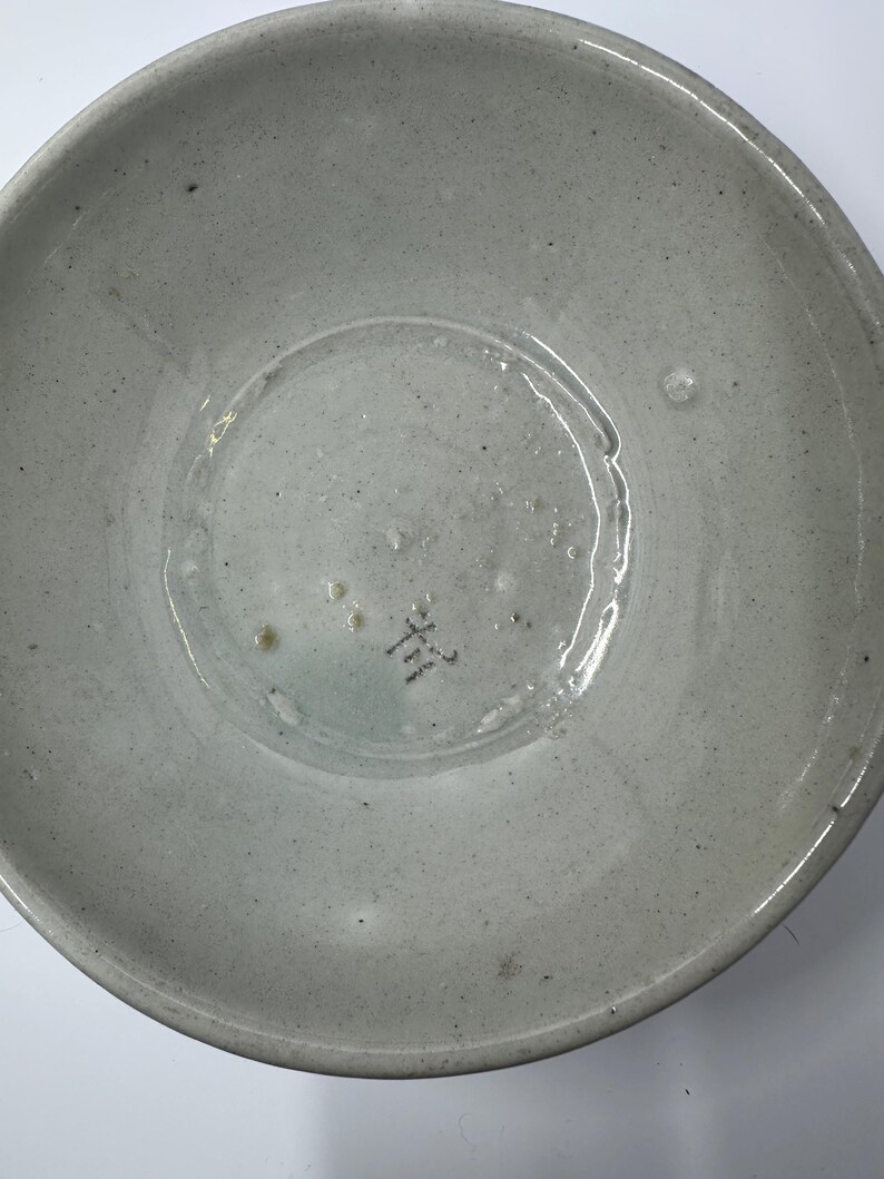 May include: A light gray ceramic bowl with a slightly textured surface. The bowl has a wide, shallow design with a rounded base and a slightly raised rim. The bottom of the bowl has a small, dark marking.