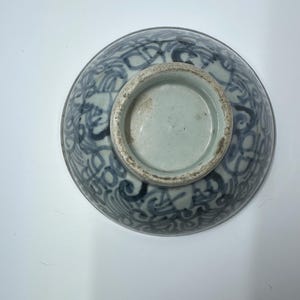 May include: A blue and white ceramic bowl with a decorative pattern. The bowl's exterior features a detailed, swirling design in shades of blue against a white background. The base of the bowl is unglazed and shows a light blue interior.