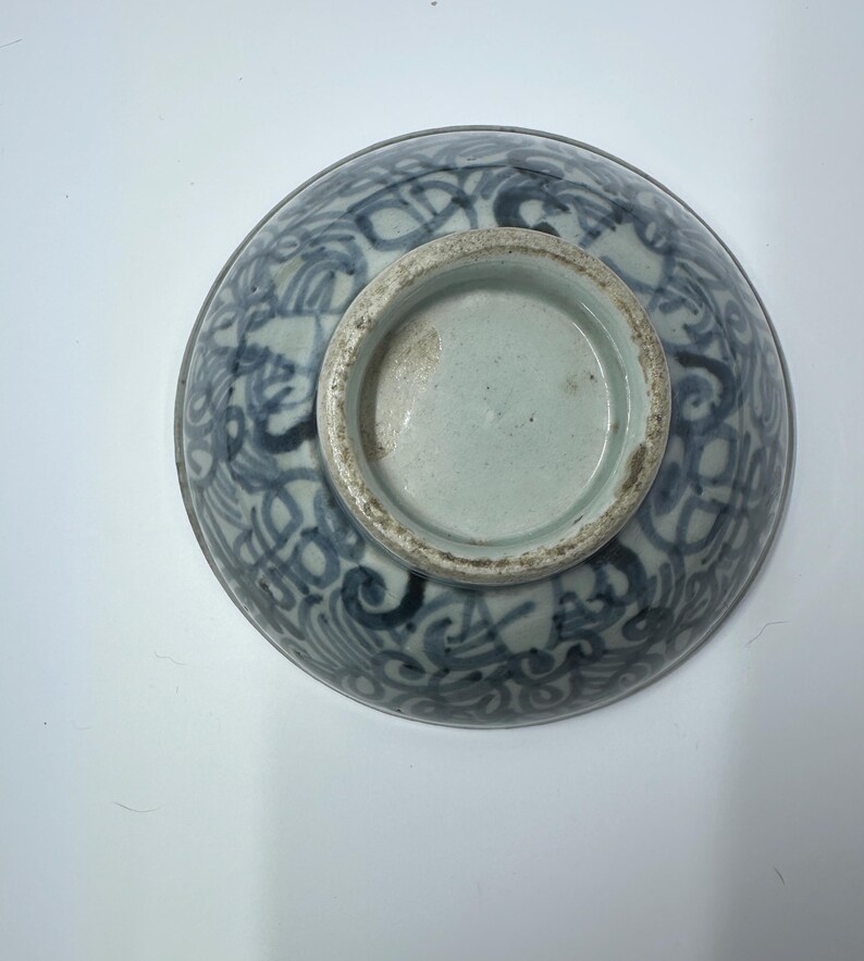 May include: A blue and white ceramic bowl with a decorative pattern. The bowl's exterior features a detailed, swirling design in shades of blue against a white background. The base of the bowl is unglazed and shows a light blue interior.