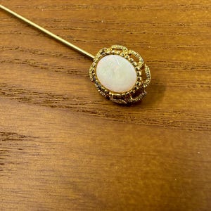 Puede incluir: Un prendedor de sombrero dorado con una piedra ovalada blanquecina engastada en un marco decorativo y texturizado. El prendedor está sobre una superficie de madera, mostrando el diseño de la joya.