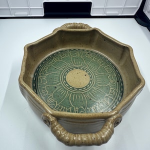 Puede incluir: Un plato de cerámica hecho a mano con forma octogonal y dos asas. El plato presenta un exterior marrón claro y un diseño floral verde y beige en el interior. Las asas texturizadas añaden a la estética rústica.