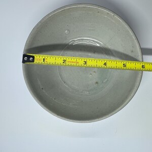 May include: A light gray ceramic bowl with a 6-inch diameter is measured with a yellow tape measure. The bowl has a simple, round design and a slightly textured surface. The bowl is likely for serving food.
