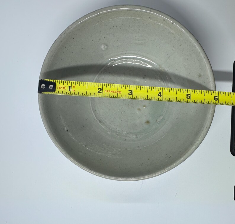 May include: A light gray ceramic bowl with a 6-inch diameter is measured with a yellow tape measure. The bowl has a simple, round design and a slightly textured surface. The bowl is likely for serving food.