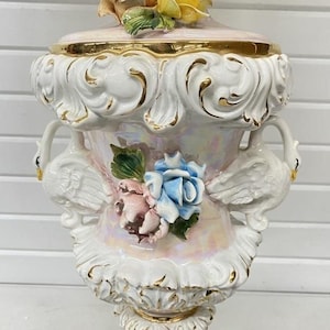 May include: A decorative ceramic lamp with a white base and a pink and gold-trimmed top. The lamp features swan handles and is adorned with floral and scrollwork details. The lamp has a gold base and is decorated with pink, blue, and yellow flowers.