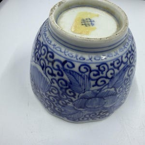 May include: A blue and white ceramic bowl turned upside down, featuring intricate floral patterns and swirling scrollwork designs. The base of the bowl displays a small, rectangular yellow sticker with a blue grid-like symbol printed on it.