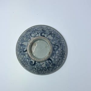 May include: A ceramic bowl with a blue and white floral pattern. The bowl is round with a footed base. The design features swirling lines and shapes. The bowl is viewed from the bottom.