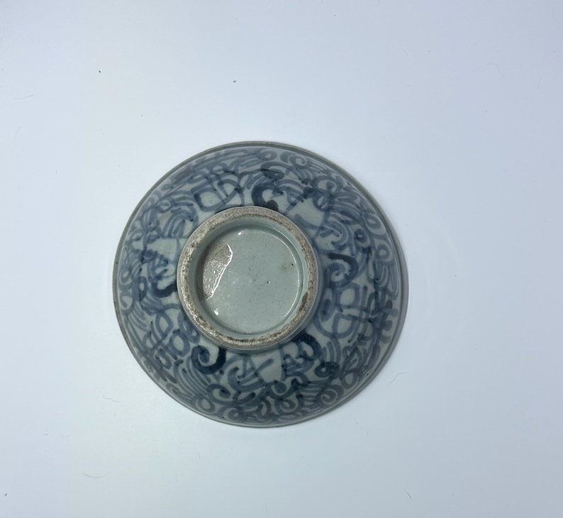 May include: A ceramic bowl with a blue and white floral pattern. The bowl is round with a footed base. The design features swirling lines and shapes. The bowl is viewed from the bottom.