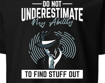 Op de afbeelding: Zwart T-shirt met witte tekst die " onderschat mijn vermogen om dingen uit te vinden niet ". Een cartoonafbeelding van een persoon met een fedora en een pak bevindt zich in het midden van de tekst.