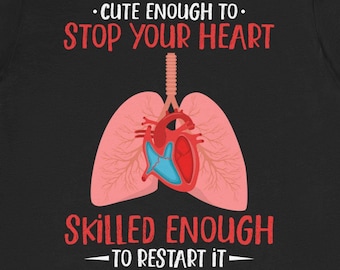 Può includere: Maglietta nera con una grafica di un cuore e polmoni. Il testo recita "Abbastanza carino da fermare il tuo cuore. Abbastanza abile da riavviarlo."