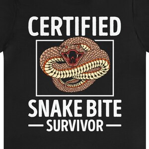 May include: Black t-shirt with white text that reads "CERTIFIED SNAKE BITE -SURVIVOR-". A graphic of a brown snake with red markings is in the center of the shirt.