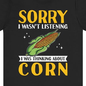 Op de afbeelding: Zwart T-shirt met een gele afbeelding van een maïskolf en de tekst "Sorry, ik luisterde niet, ik dacht aan maïs".