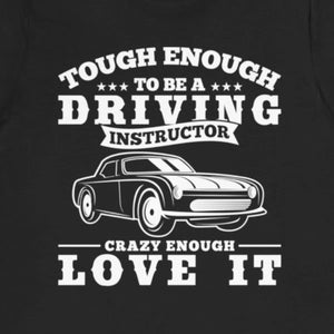 Op de afbeelding: Zwart T-shirt met witte tekst die luidt: "Tough Enough to be a Driving Instructor. Crazy Enough Love It."  Een zwart-witte illustratie van een klassieke auto bevindt zich in het midden van het shirt.