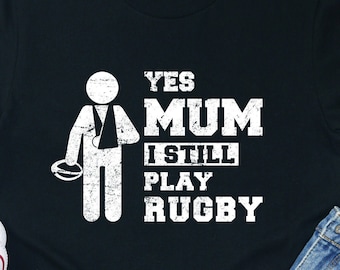 May include: Black t-shirt with a white graphic of a stick figure with a broken arm holding a rugby ball. The text "Yes Mum I Still Play Rugby" is printed on the shirt.