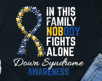 Op de afbeelding: Zwart T-shirt met een blauw en geel lint gemaakt van harten. De tekst "In deze familie vecht niemand alleen. Down syndroom bewustzijn" staat op het T-shirt gedrukt.