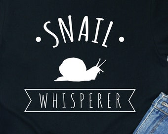 May include: Black t-shirt with white text that reads "Snail Whisperer" and a white silhouette of a snail. The shirt is laid out on a wooden surface with a pair of white sneakers and denim shorts.