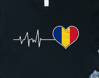 May include: Black t-shirt with a white heartbeat graphic and a heart-shaped Romanian flag. The flag is yellow, blue, and red. The shirt is on a white wooden surface with a pair of white sneakers and blue denim shorts.