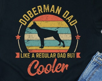 Op de afbeelding: Zwart T-shirt met een vintage-stijl grafische afbeelding van een Dobermann Pinscher silhouet voor een zonsondergang. De tekst "Doberman Dad" staat boven de afbeelding en "Like a regular dad but cooler" staat onder de afbeelding.