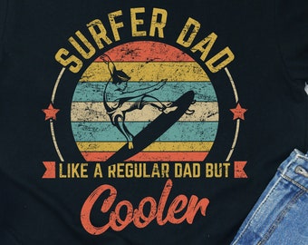 Può includere: Maglietta nera con una grafica in stile vintage che dice "Surfer Dad Like a Regular Dad But Cooler". La grafica presenta un surfista che cavalca un'onda con uno sfondo di tramonto.