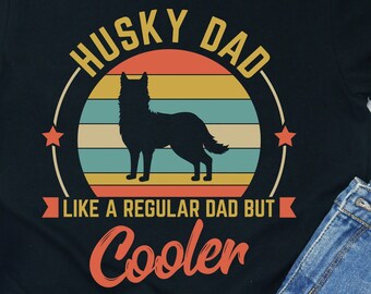 May include: Black t-shirt with a graphic that reads "Husky Dad" above a silhouette of a husky dog. The text "Like a regular dad but cooler" is below the dog. The shirt is paired with denim shorts and white sneakers.