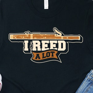 Puede incluir: Camiseta negra con una ilustración marrón y blanca de un fagot y el texto "I reed a lot".