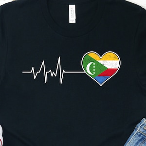 May include: Black t-shirt with a white heartbeat graphic and a heart-shaped Comoros flag. The flag is yellow, green, blue, and white.