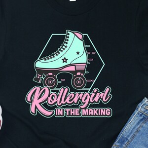 Może przedstawiać: Czarny T-shirt z grafiką przedstawiającą turkusowe rolki z różowymi akcentami i tekstem "Rollergirl in the making".