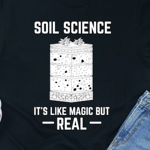 Peut inclure: T-shirt noir avec du texte blanc qui dit "Soil Science It's like magic but -REAL-" et un graphique d'un profil de sol.