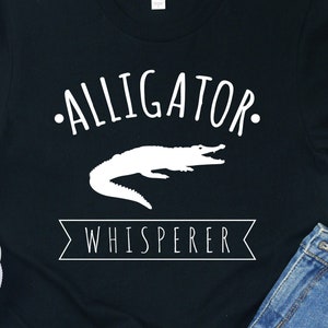 May include: Black t-shirt with a white alligator graphic and the text "Alligator Whisperer".